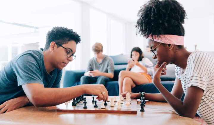 Young friends playing chess and others using smartphone