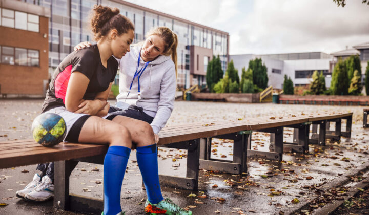 Young rugby player sitting on a bench outside a school with their head down looking sad next to rugby ball with their coaches arm over them trying to comfort them