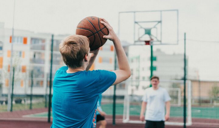 Young British basketball player wearing a blue shirt shooting basketball shot with team mate wearing white shirt in the guarding the net