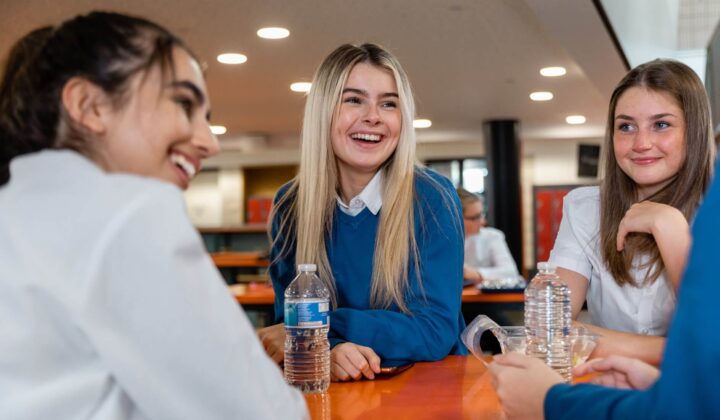 An over the shoulder view of a group of best friends catching up in the dinner hall of a high school in England. They are chatting together and smiling and laughing.