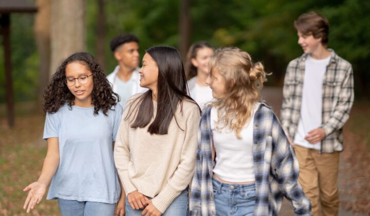 A small group of teenagers walk casually outside on a fall day together as they laugh and talk amongst themselves. They are each dressed casually in warm layers and enjoying hanging out together.