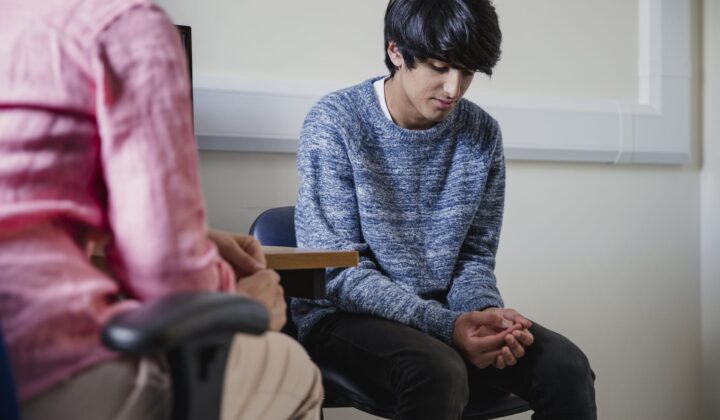 Young boy sitting on chair looking down with hands together whilst talking to person about their mental health